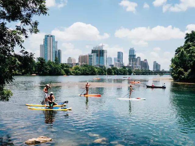 A Landscape in Austin showing many small boat riders on a lake with many trees behind them, followed by skyscrapers